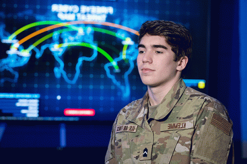 Male student with short, dark hair, and wearing military fatigues with an ‘Air Force’ patch, stands looking left to right in a darkly lit classroom with a bright, digital world map visible in the background. 